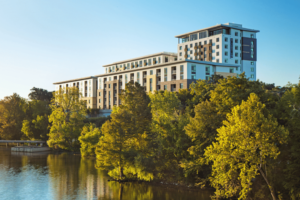 Diagonal elevation of Berkshire Riverview, a six‑story mixed‑use residential complex along Lady Bird Lake. The structure features a cast‑in‑place concrete podium at lower levels with steel‑framed upper floors, accented by brick and fiber‑cement panels, recessed balconies, and a consistent grid of structural columns and slab edges.